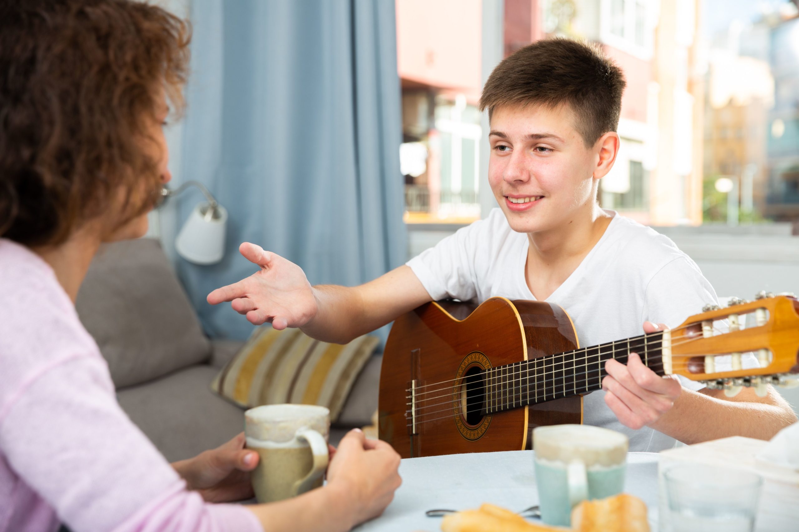 Teenager with guitar talking to mother - How to Support Your Child This School Year—Without Pushing Them to Be Perfect: Be a Gardener, Not a Carpenter - Cheerful teenage boy with guitar friendly talking to his mother at home table..